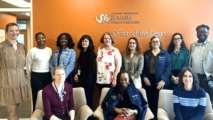 A group of twelve people standing and sitting in front of the Office of the Dean sign at Drexel University's Dornsife School of Public Health
