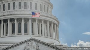 The American flag waves in front of the U.S. Capitol building, featuring its distinctive dome and classical columns against a partly cloudy sky