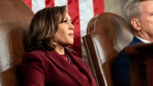 Close up of Kamala Harris in a maroon suit jacket sitting near the US flag