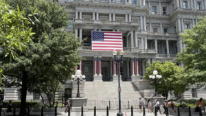 Historic government building with an American flag and patriotic banners, surrounded by trees and street lamps, with people walking by_1600x900