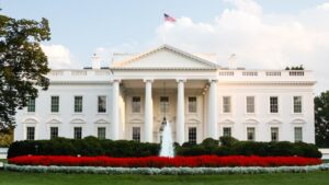 The White House North Portico with an American flag flying on top, framed by blue skies, lush greenery, and a red flower bed in the foreground